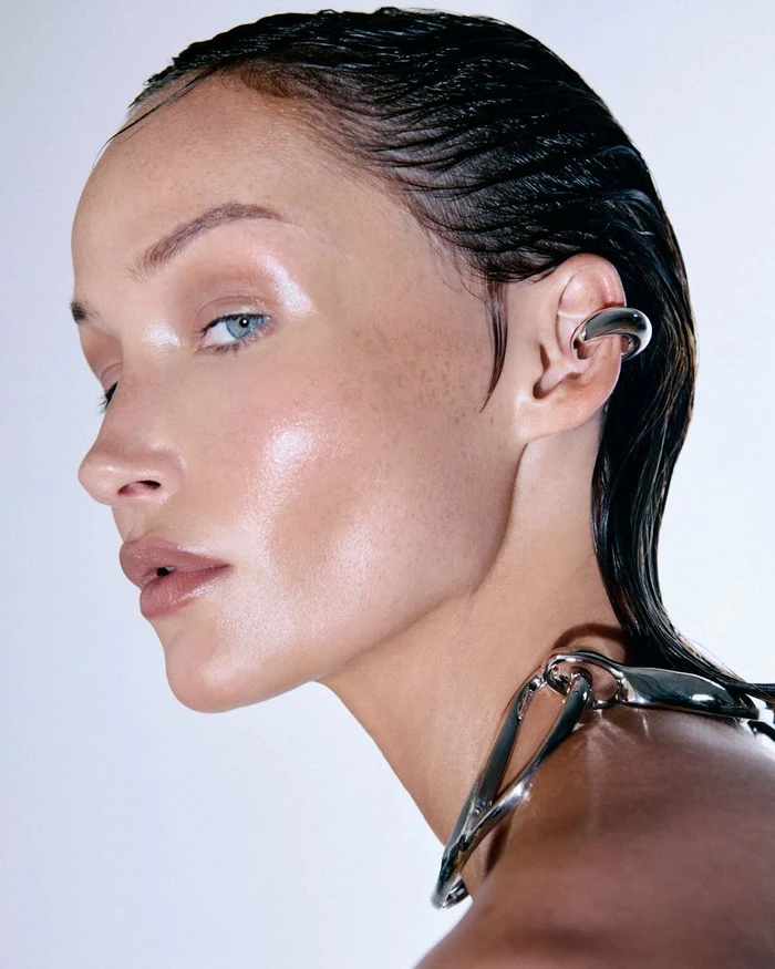 Close-up profile of a woman with slicked-back wet-look hair wearing a sculptural silver ear cuff and metal collar against a white background.