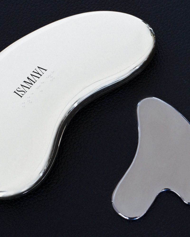 Close-up view of two polished metallic chrome Isamaya gua sha facial sculpting tools resting on a black textured leather surface.