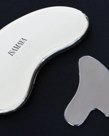 Close-up view of two polished metallic chrome Isamaya gua sha facial sculpting tools resting on a black textured leather surface.