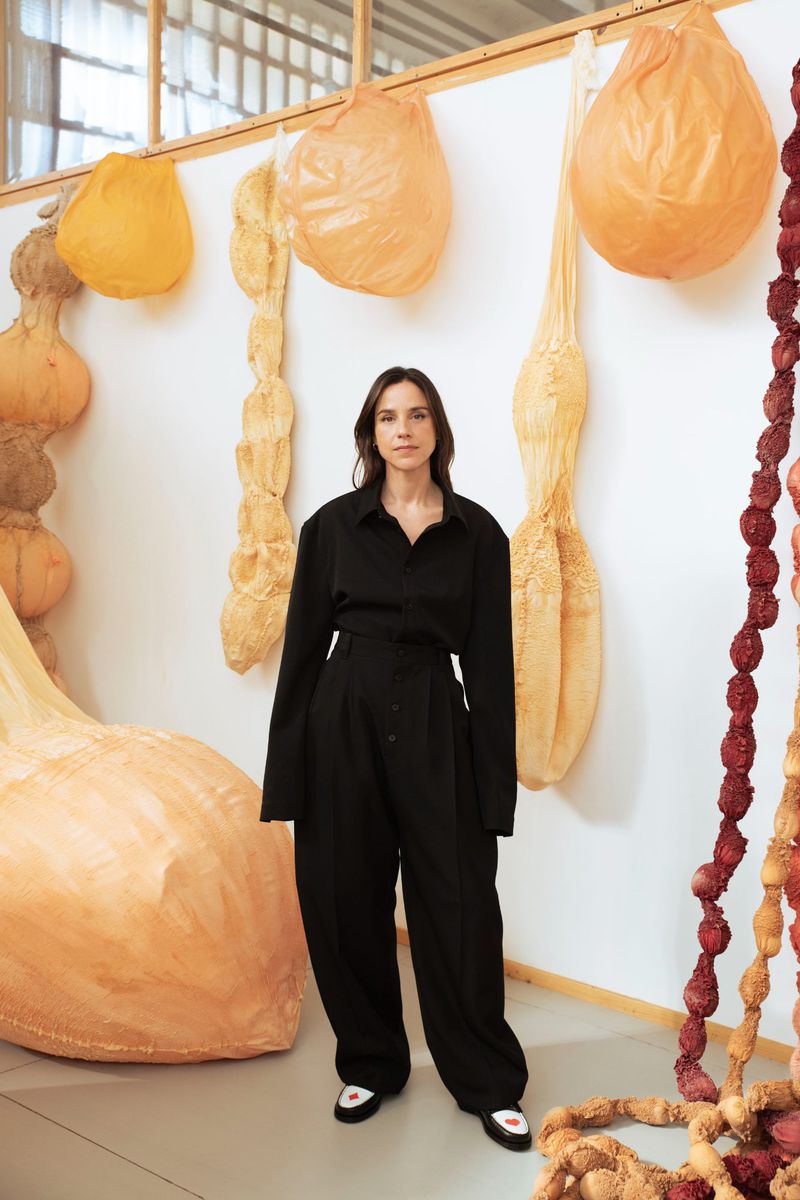 Adult woman in a black jumpsuit stands in front of various beige and red hanging sculptures against a white wall in a bright space.