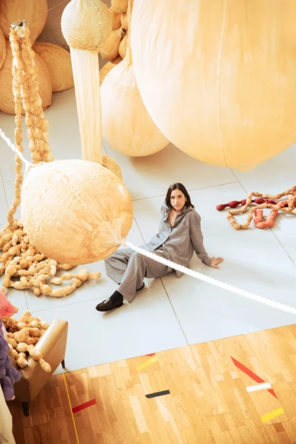 Woman in grey loungewear sits amidst oversized, beige, textured sculptural elements in an indoor setting with warm, wood-paneled flooring.