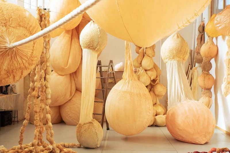 Surreal indoor art installation featuring multiple large, beige, balloon-like shapes hanging from the ceiling in a brightly lit room.