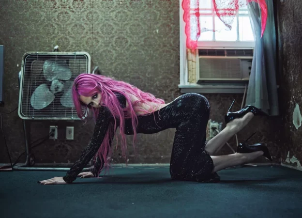 Woman with long pink hair crawling in a sparkling black backless gown and stiletto heels inside a vintage room.