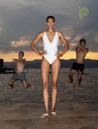 Model in a white one-piece swimsuit standing on a coastal dock at sunset with two boys jumping into the water behind her.