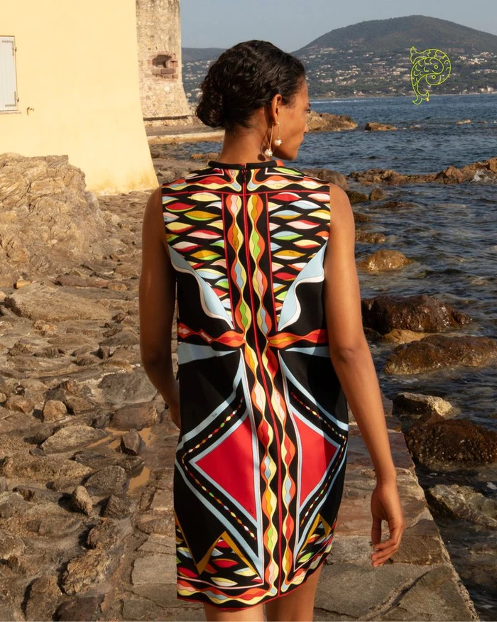 Model Mar G seen from behind wearing a colorful geometric print sleeveless dress along a rocky Mediterranean coast.
