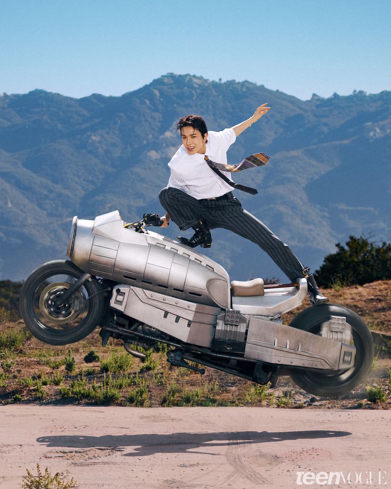 Man dynamic posing atop a futuristic silver motorcycle hovering in mid-air against mountain hills under a clear blue sky.