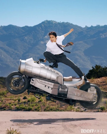 Man dynamic posing atop a futuristic silver motorcycle hovering in mid-air against mountain hills under a clear blue sky.