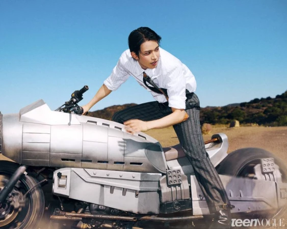 Man in a white shirt and striped trousers riding a futuristic metallic motorcycle against a bright blue sky.