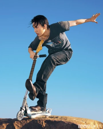 Man in a graphic t-shirt and jeans balancing dynamically on a kick scooter holding an electric guitar against a clear blue sky.