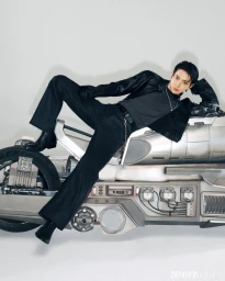 Man in a black leather jacket and dark trousers reclining across a futuristic metallic motorcycle against a studio backdrop.