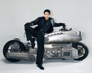 Man in an all-black leather outfit sitting atop a futuristic metallic motorcycle prop against a white studio background.