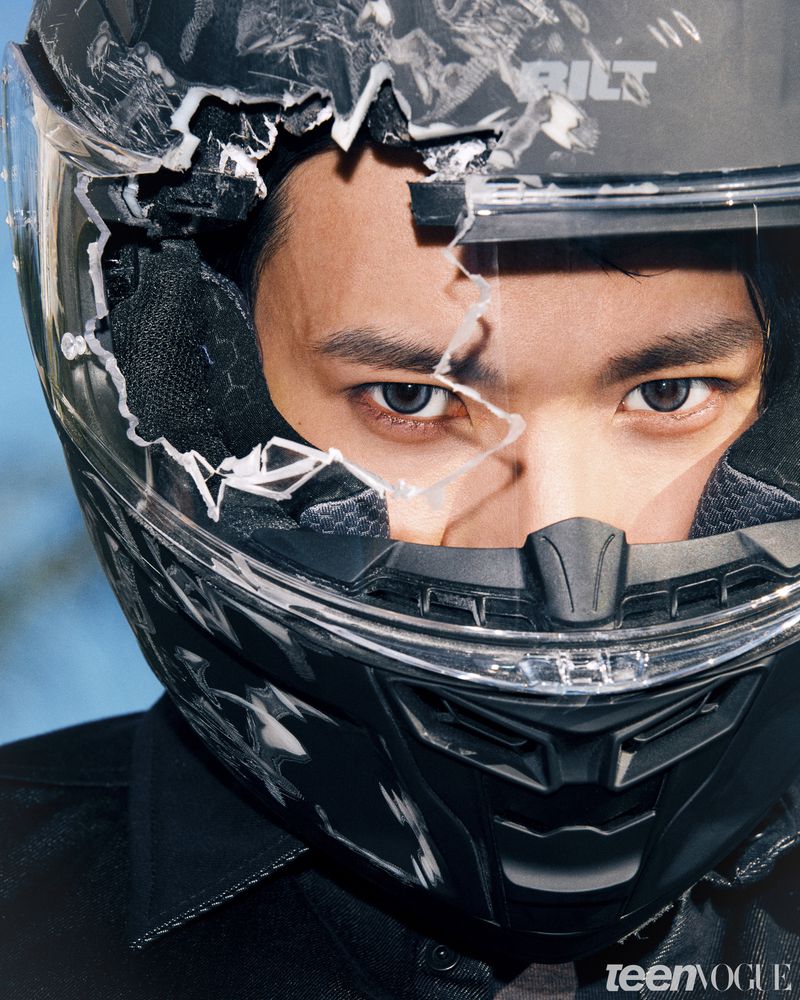 Close-up portrait of an Asian man wearing a scuffed black motorcycle helmet with a shattered visor revealing his eyes.