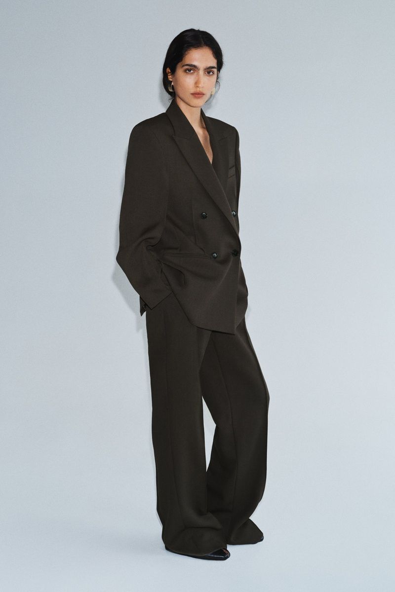 Model wearing an oversized dark tailored double-breasted pantsuit standing against a seamless light studio backdrop.