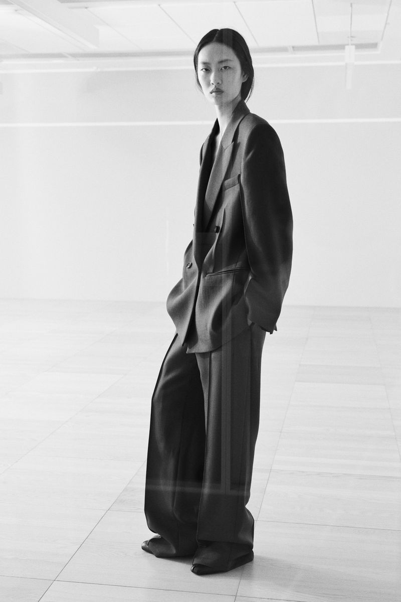 Model Ruiyi Zhang poses in an oversized dark tailored pantsuit in a bright minimalist interior space.