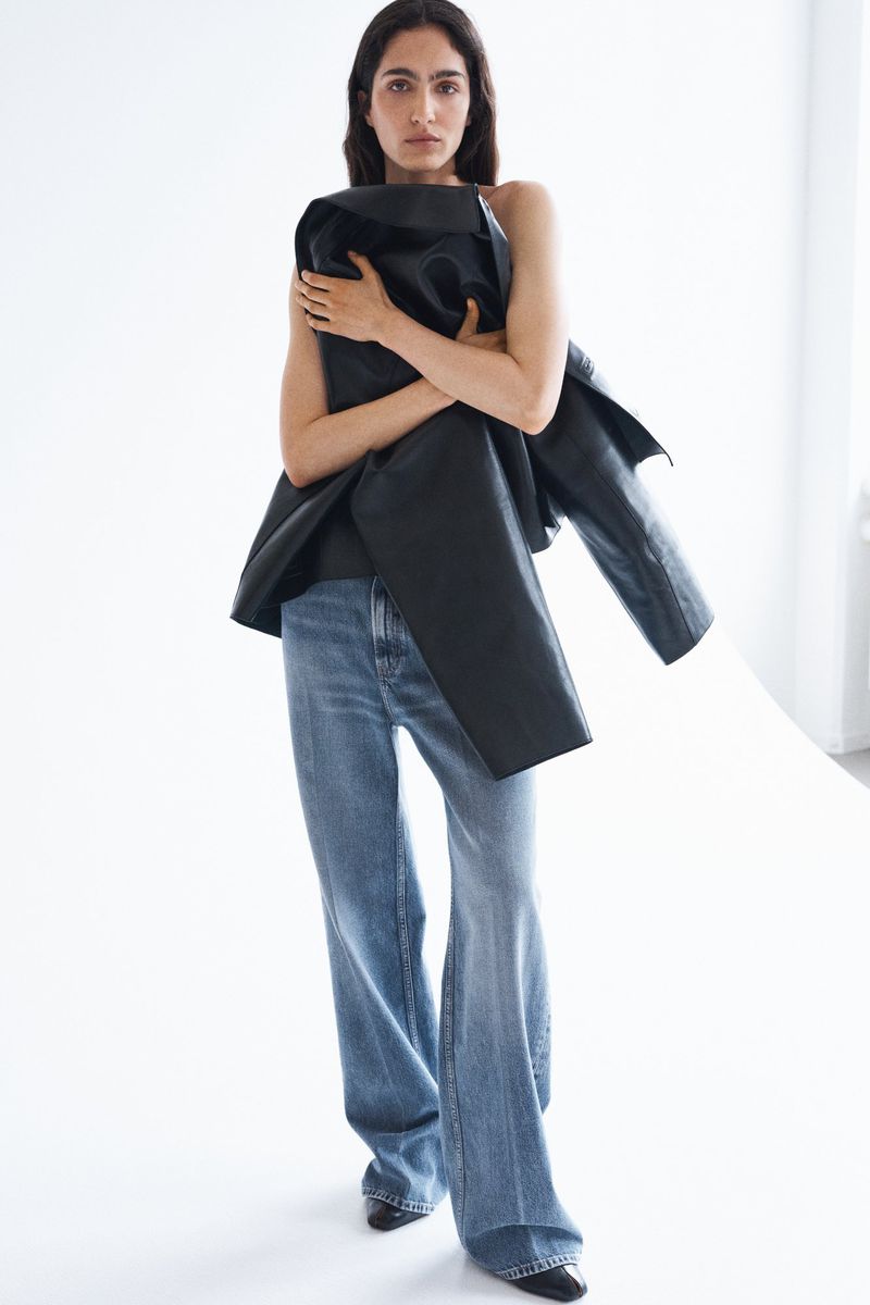 Model wearing wide-leg blue denim jeans and holding a black leather jacket against a bright white seamless background.