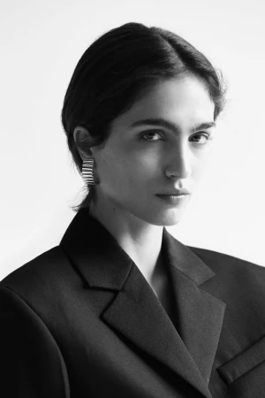 Close-up portrait of model Sara Caballero in a black tailored blazer and ribbed metallic cuff earring against a white background.