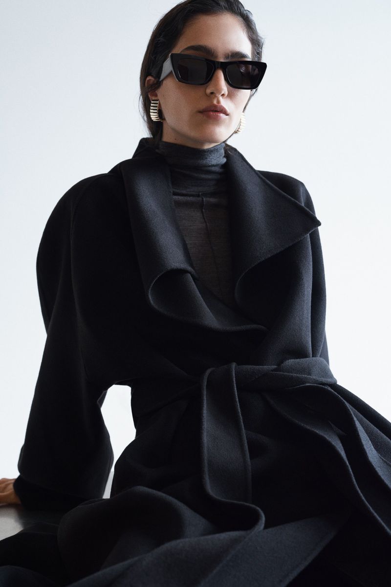 Model wearing dark sunglasses and an oversized black belted wrap coat against a white background.