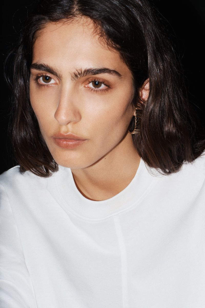Close-up portrait of model Sara Caballero wearing a white crew neck top with gold earrings against a black background.