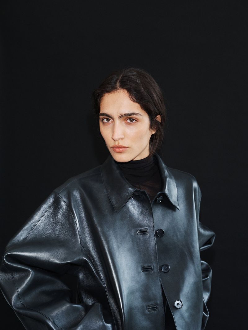 Model wearing an oversized black leather jacket over a black turtleneck against a dark seamless studio backdrop.