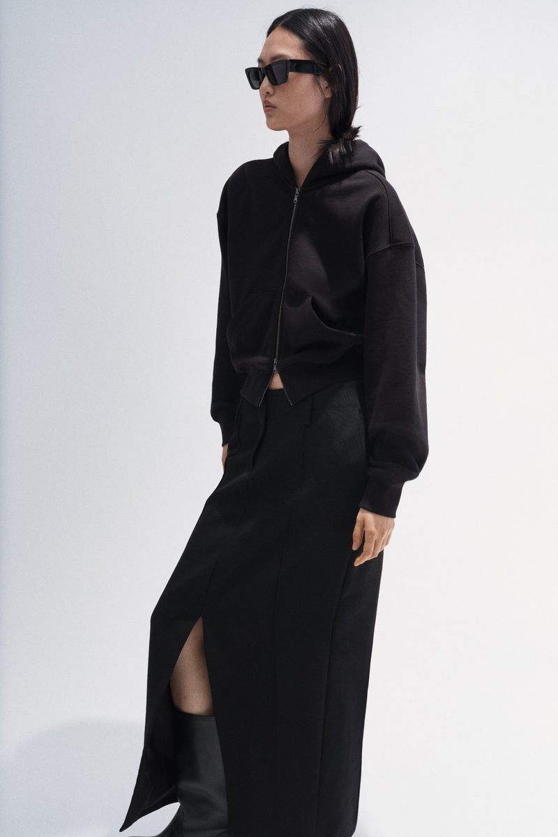 East Asian model in black sunglasses, a black zip-up hoodie, and a matching slit maxi skirt against a white studio backdrop.