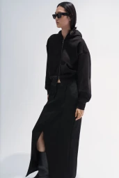 East Asian model in black sunglasses, a black zip-up hoodie, and a matching slit maxi skirt against a white studio backdrop.