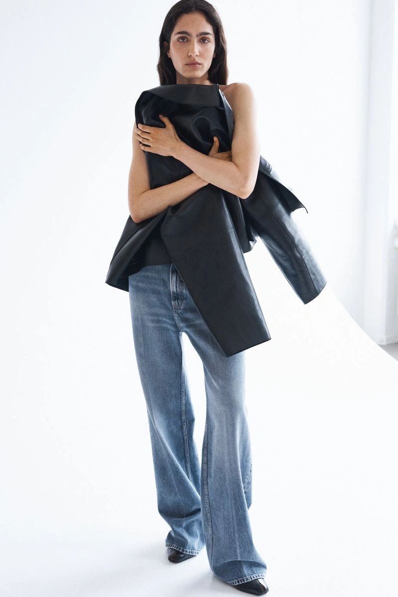 Model clutching a black leather jacket to her chest wearing wide-leg light blue jeans against a white studio backdrop.