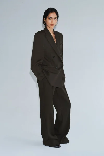 Model Sara Caballero wearing an oversized dark brown tailored trouser suit against a pale studio backdrop.