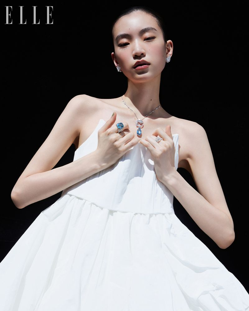 East Asian woman in a white dress wearing a blue gemstone necklace and rings against a black background.