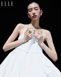 East Asian woman in a white dress wearing a blue gemstone necklace and rings against a black background.