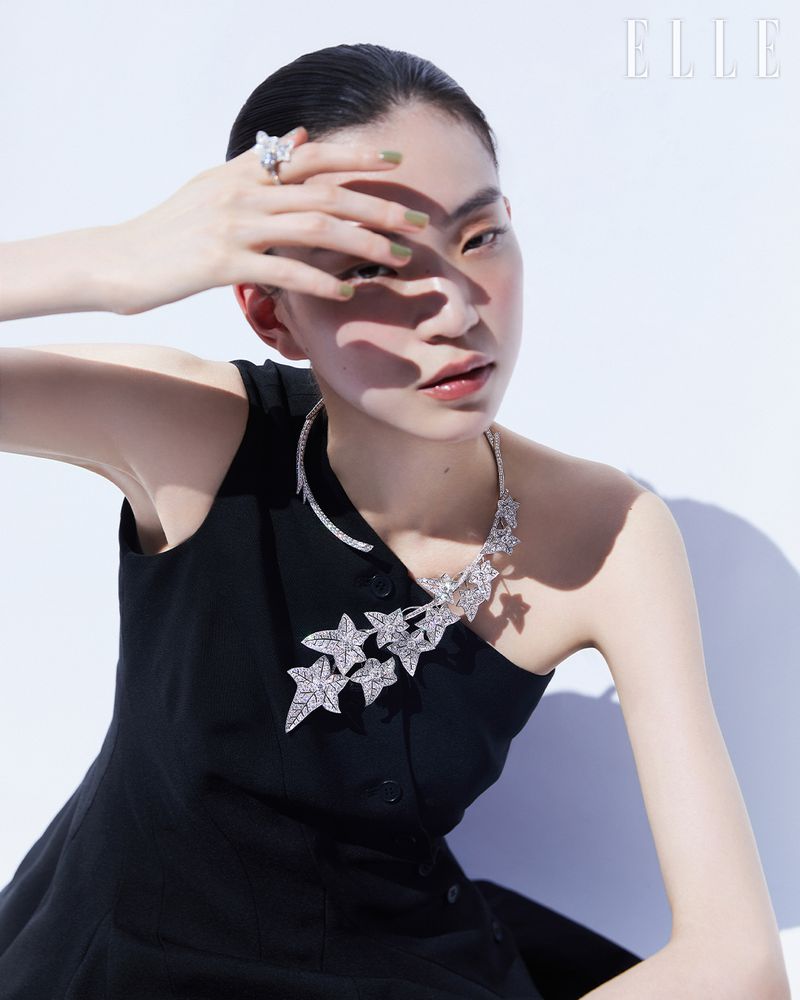 An East Asian woman in a black asymmetrical dress wearing an intricate diamond leaf necklace and ring against a white studio background.