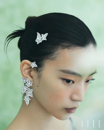 Close-up portrait of an East Asian woman with slicked hair wearing diamond leaf-shaped earrings and hair jewelry against a green backdrop.