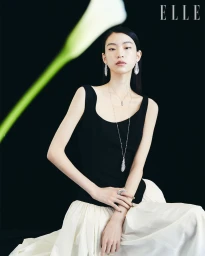 East Asian model in a black scoop-neck top and white pleated skirt wearing fine diamond jewellery against a black background.
