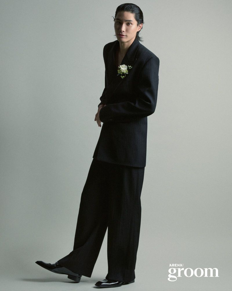 Model Jinyeong Pak wearing an oversized black suit with a white floral boutonniere against a neutral grey studio background.