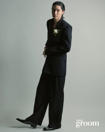 Model Jinyeong Pak wearing an oversized black suit with a white floral boutonniere against a neutral grey studio background.