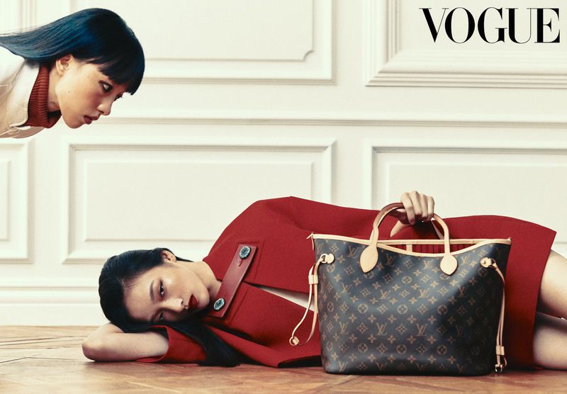 Model lying on a wooden floor in a red coat holding a monogram tote bag as another model looks down against a white paneled wall.