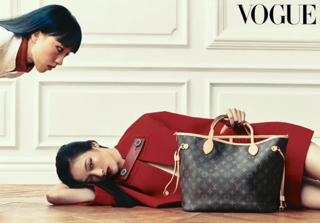 Model lying on a wooden floor in a red coat holding a monogram tote bag as another model looks down against a white paneled wall.