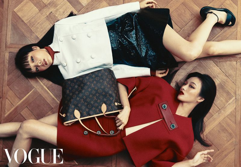 Two fashion models lying on a herringbone parquet wood floor in red and white structured coats beside a monogram tote bag.