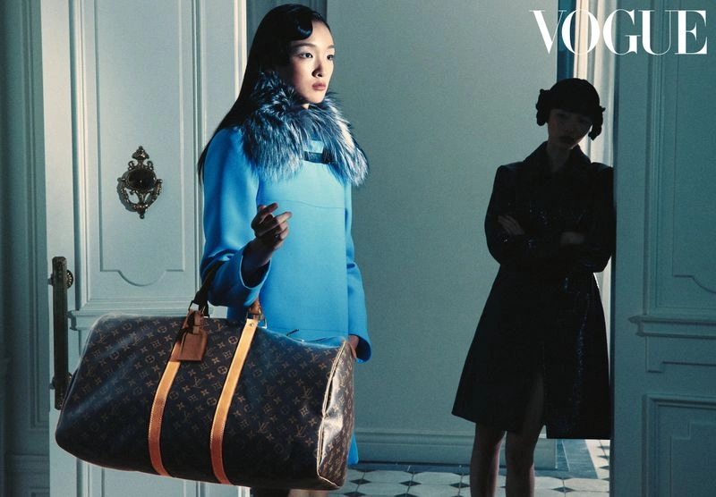 Model Shiqi Fang in a vibrant blue coat holding a large brown monogram duffle bag by an interior doorway.