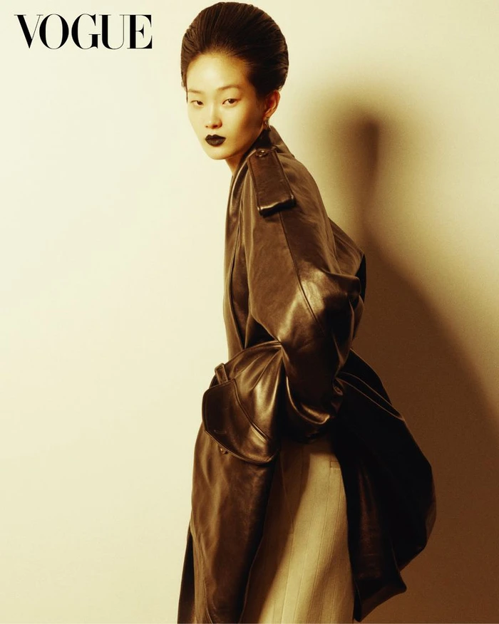 Model Hyunji Shin posing in an oversized dark leather trench coat and bold dark lipstick against a warm studio background.