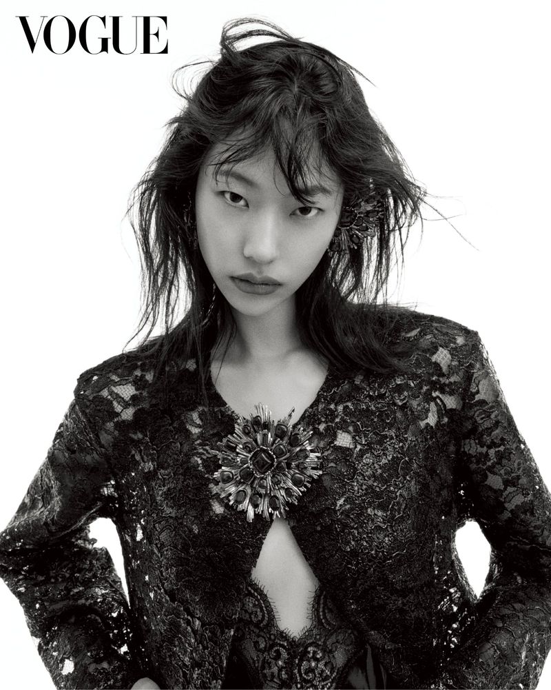 East Asian female model with tousled dark hair wearing a black lace jacket with an ornate brooch against a white backdrop.