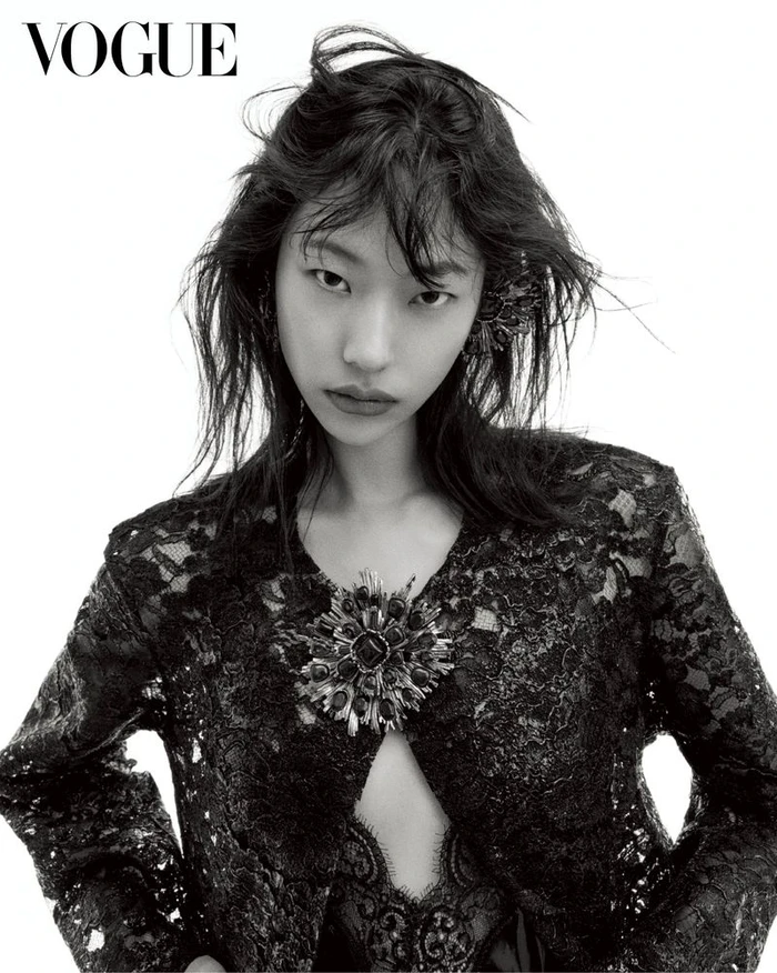 East Asian female model with tousled dark hair wearing a black lace jacket with an ornate brooch against a white backdrop.