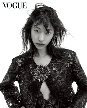East Asian female model with tousled dark hair wearing a black lace jacket with an ornate brooch against a white backdrop.