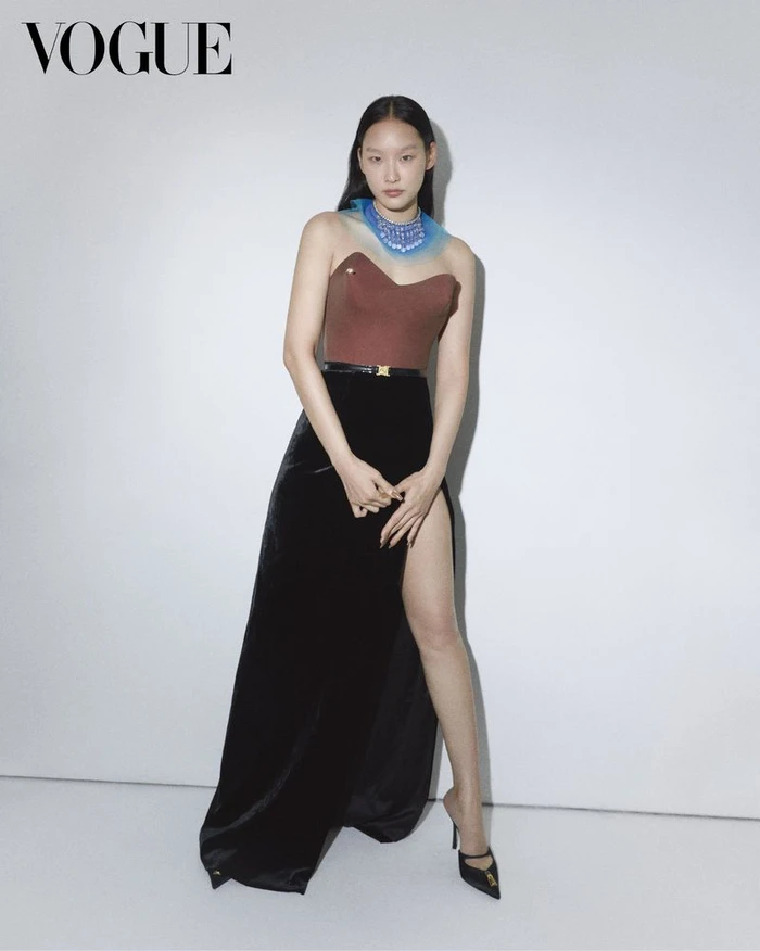 East Asian female model in a brown strapless corset top and high-slit black velvet skirt against a white studio background.