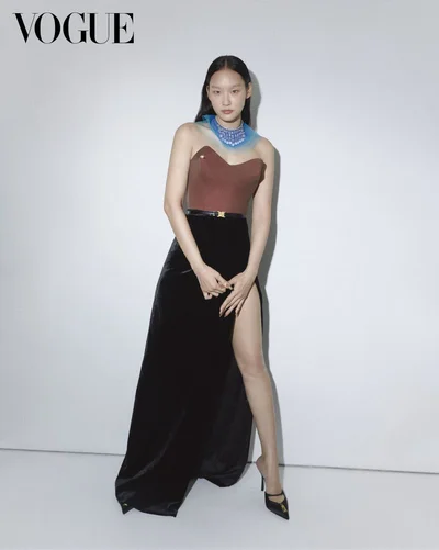 East Asian female model in a brown strapless corset top and high-slit black velvet skirt against a white studio background.