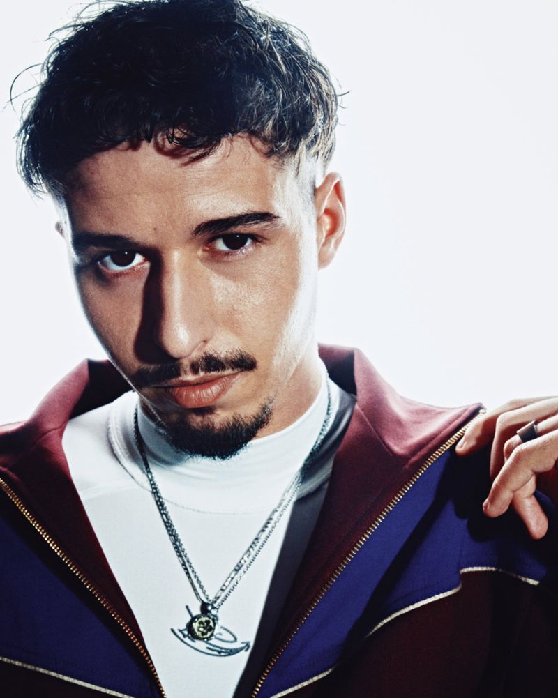 Close-up portrait of a man with a goatee wearing a burgundy and purple track jacket over a white top against a white background.