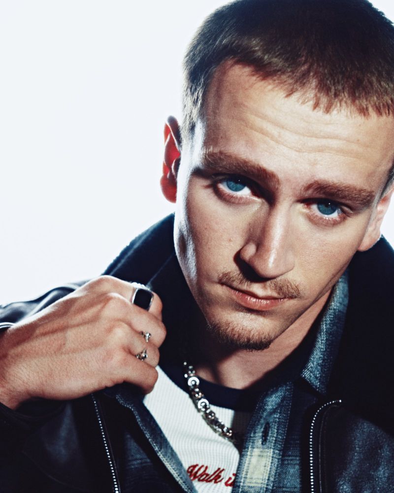 Close-up portrait of a young blue-eyed man in a black leather jacket and silver rings against a bright white background.