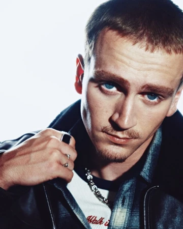 Close-up portrait of a young blue-eyed man in a black leather jacket and silver rings against a bright white background.