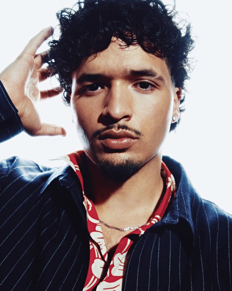 Close-up portrait of a man with curly hair wearing a dark pinstripe shirt over a red floral collar against a white background.