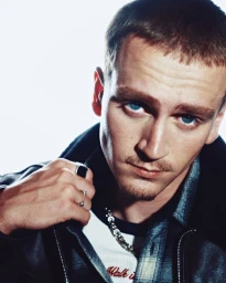 Close-up portrait of a young blue-eyed man in a black leather jacket and silver rings against a bright white background.