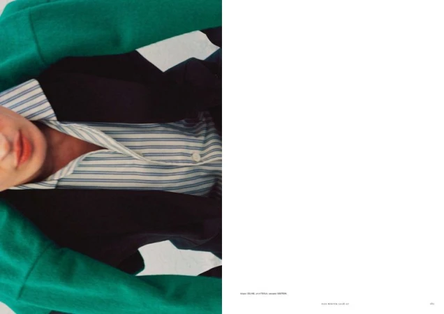 Close-up of a model wearing an emerald green coat, black blazer, and blue striped collared shirt beside white negative space.